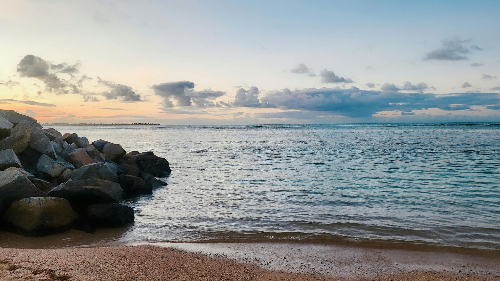 Calm coastal shoreline with gentle waves lapping a sandy beach, large rocks along the shore, and a wide horizon under a softly clouded sky.