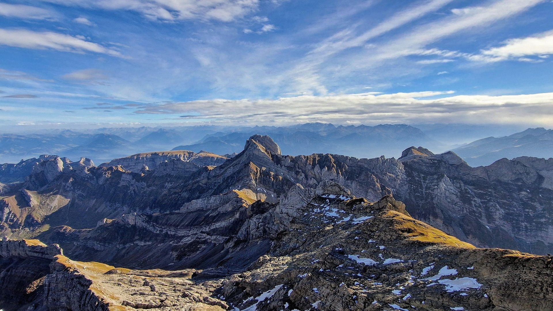 Rocky mountain habitat with jagged peaks, steep ridges, and patches of snow under a wide blue sky with scattered clouds.