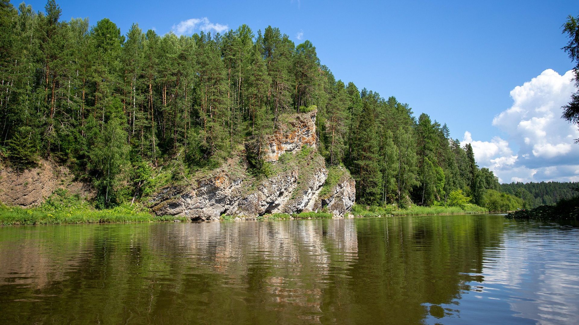 A calm river flowing through a taiga forest, with dense coniferous trees lining the banks and rocky cliffs reflected in the water under a clear blue sky.